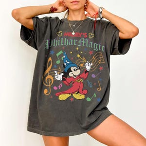 May include: Dark gray t-shirt featuring Mickey Mouse as a conductor with the text "Mickey's PhilharMagic". The graphic includes musical notes, stars, and a colorful design. The shirt has short sleeves and a relaxed fit.