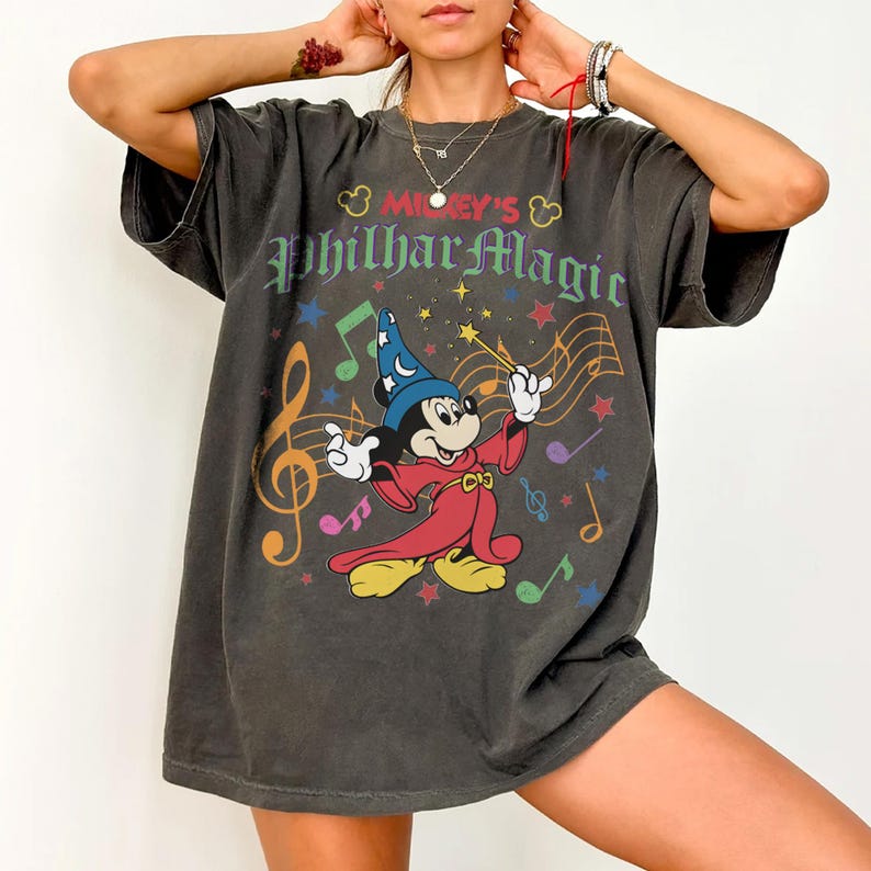 May include: Dark gray t-shirt featuring Mickey Mouse as a conductor with the text "Mickey's PhilharMagic". The graphic includes musical notes, stars, and a colorful design. The shirt has short sleeves and a relaxed fit.