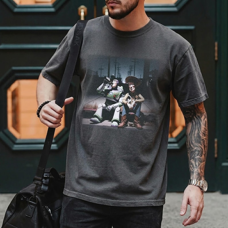 Puede incluir: Camiseta gris oscuro con un gr&aacute;fico de Buzz Lightyear y Woody de Toy Story. La imagen muestra a los personajes juntos en una escena callejera. La camiseta tiene un aspecto vintage y desgastado.