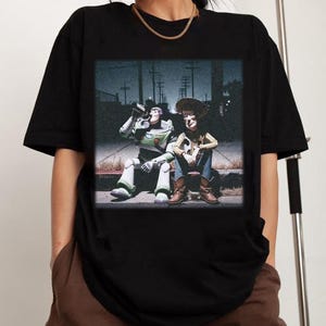 Puede incluir: Camiseta negra con un gr&aacute;fico de Buzz Lightyear y Woody de Toy Story sentados juntos. Buzz est&aacute; bebiendo de una lata. La imagen tiene un aspecto vintage, ligeramente desgastado.