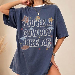 May include: A dark blue short-sleeve t-shirt featuring the text "YOU'RE A COWBOY LIKE ME" in a white western-style font. The graphic includes illustrations of brown cowboy boots, a hat, a yellow star, a planet, clouds, and a small rocket ship.