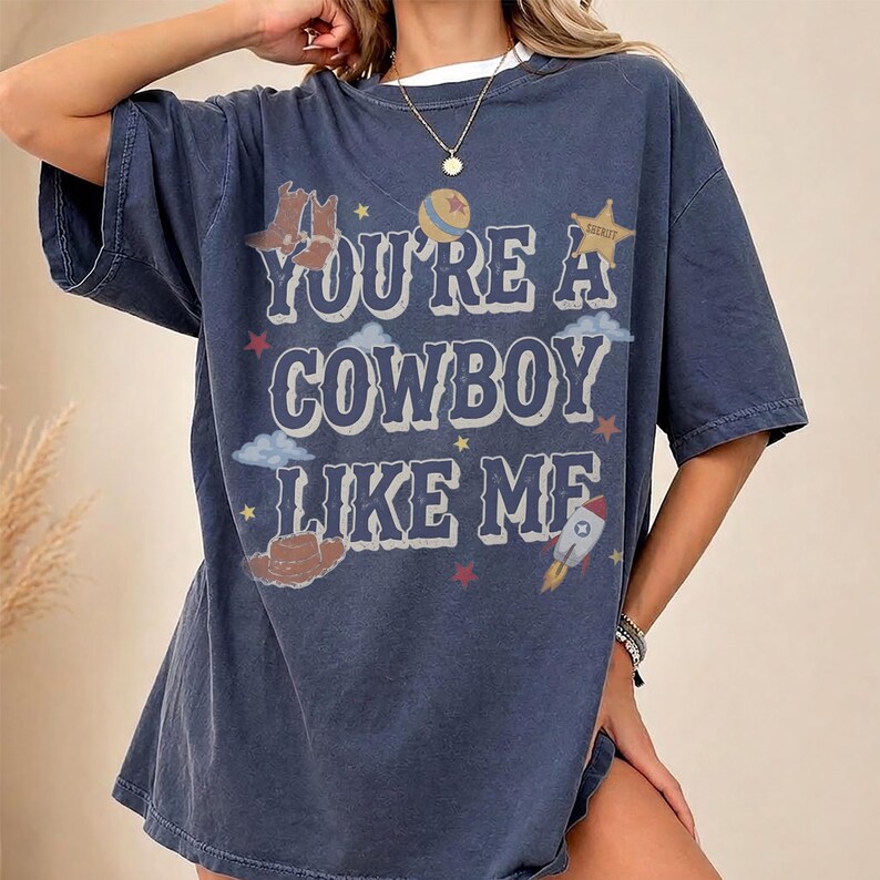 May include: A dark blue short-sleeve t-shirt featuring the text "YOU'RE A COWBOY LIKE ME" in a white western-style font. The graphic includes illustrations of brown cowboy boots, a hat, a yellow star, a planet, clouds, and a small rocket ship.