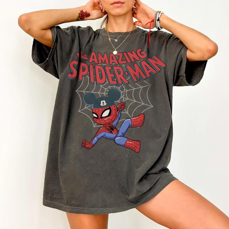 May include: Dark gray t-shirt featuring a cartoon Spider-Man character wearing a Mickey Mouse hat, hanging from a web. The text "THE AMAZING SPIDER-MAN" is printed across the top in red and blue. The shirt has short sleeves.