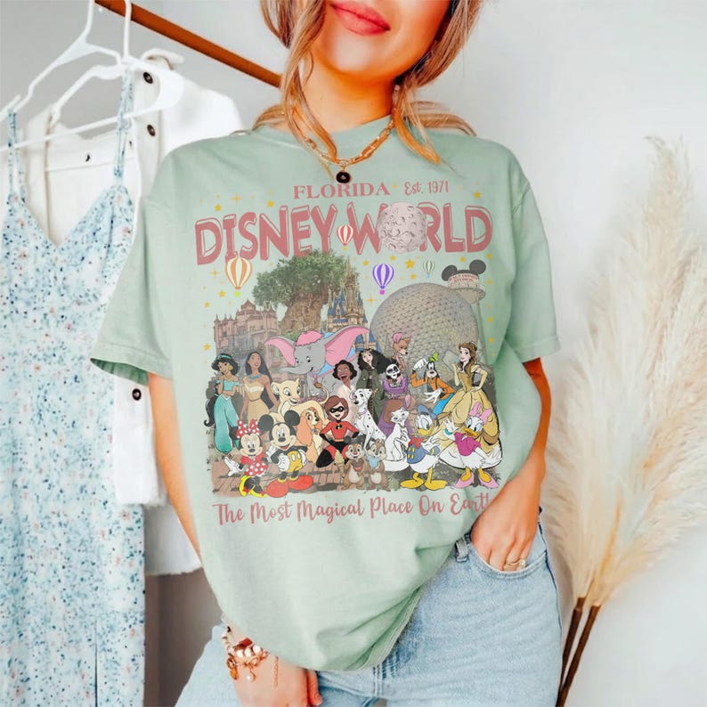May include: A light green t-shirt with a graphic featuring Disney World characters and the text "FLORIDA Est. 1971 DISNEY WORLD The Most Magical Place On Earth". The design includes various Disney characters and park icons.