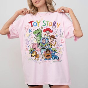 Puede incluir: Una camiseta rosa claro con un gr&aacute;fico colorido de los personajes de Toy Story. Las palabras "Toy Story" est&aacute;n en rojo y azul, con garabatos y la frase "yee haw". La camiseta tiene un corte informal y de gran tama&ntilde;o.