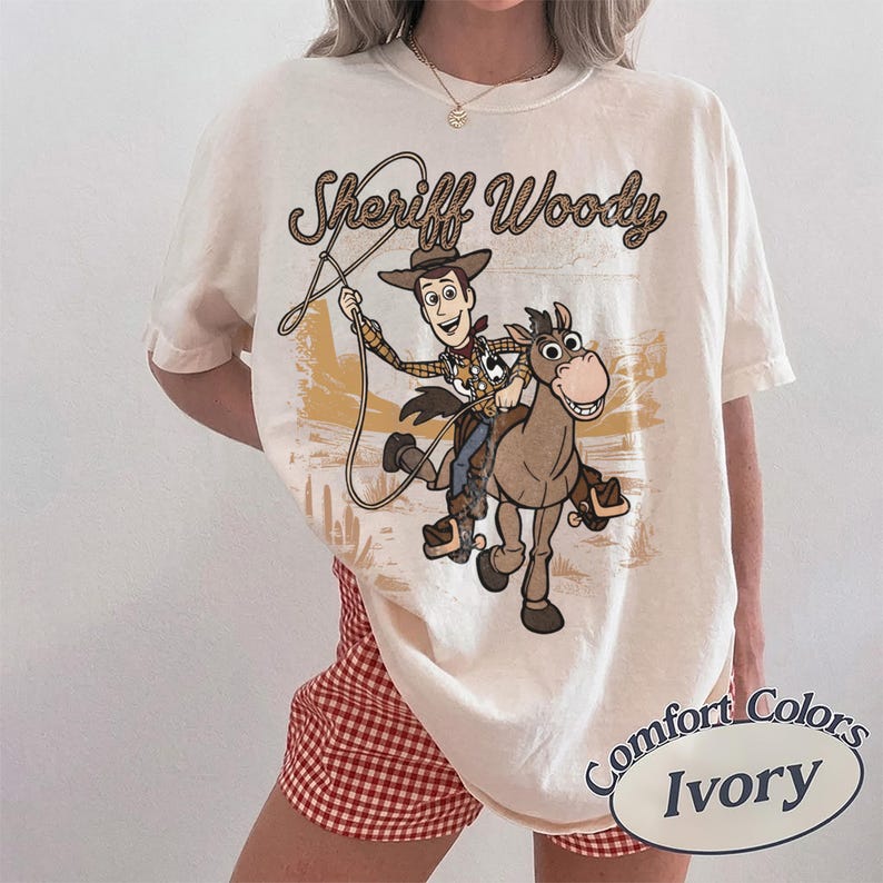 May include: An ivory t-shirt featuring a graphic print of Sheriff Woody riding a horse while holding a lasso. The text "Sheriff Woody" is printed above the character in a brown, rope-style font. The background graphic depicts a desert landscape.