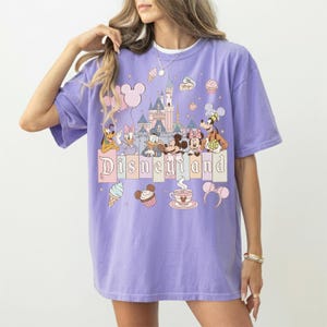 May include: A lilac t-shirt with a Disneyland graphic. The design features cartoon characters, a castle, and the word "Disneyland" in a pastel font. Additional graphics include ice cream cones, cupcakes, and Mickey Mouse ears.