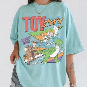 May include: A pale blue t-shirt with the words "TOY Story" in red and yellow, and the text "SINCE 1995". The shirt features a graphic of Buzz Lightyear, Woody, Bullseye, and Rex.