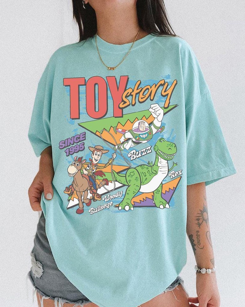 May include: A pale blue t-shirt with the words "TOY Story" in red and yellow, and the text "SINCE 1995". The shirt features a graphic of Buzz Lightyear, Woody, Bullseye, and Rex.