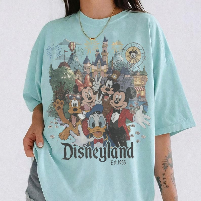 May include: A light blue t-shirt featuring a graphic print of Mickey Mouse, Minnie Mouse, Donald Duck, Goofy, and Pluto in front of a Disneyland castle. The text Disneyland Est. 1955 is printed at the bottom of the graphic in a dark font.
