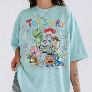 Puede incluir: Una camiseta azul claro con las palabras "TOY STORY" en letras coloridas. La camiseta muestra un gr&aacute;fico de varios personajes de la pel&iacute;cula animada, incluyendo a Woody, Buzz Lightyear y Rex, con acentos tipo garabato.