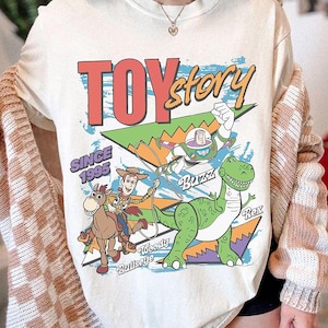May include: Cream-coloured t-shirt with a graphic design from the film "Toy Story." The design includes the text "TOY story" in red and yellow, along with illustrations of Woody, Buzz Lightyear, and Rex the dinosaur. The text "SINCE 1995" is also present.