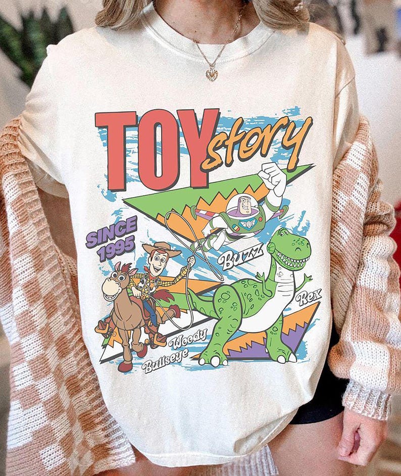 May include: Cream-coloured t-shirt with a graphic design from the film "Toy Story." The design includes the text "TOY story" in red and yellow, along with illustrations of Woody, Buzz Lightyear, and Rex the dinosaur. The text "SINCE 1995" is also present.