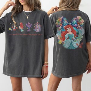 May include: Dark gray t-shirt featuring a colorful illustration of a mermaid, castle, and sea creatures. The front of the shirt has the text "I want to be where the people are". The back of the shirt has a large image of a mermaid.