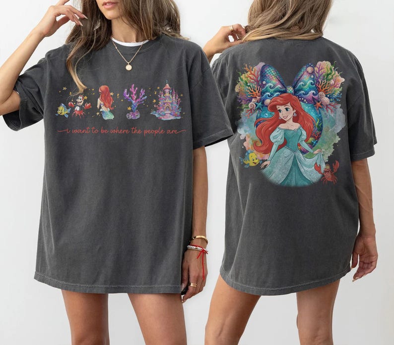 May include: Dark gray t-shirt featuring a colorful illustration of a mermaid, castle, and sea creatures. The front of the shirt has the text "I want to be where the people are". The back of the shirt has a large image of a mermaid.