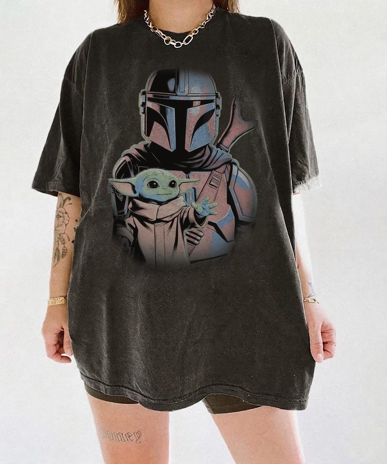 May include: A charcoal gray oversized t-shirt featuring a graphic print of the Mandalorian character wearing a helmet and armor, with the small green creature known as Grogu positioned in the foreground with one hand extended forward.