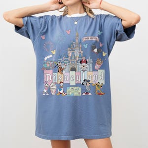 May include: Blue t-shirt featuring a colorful Disneyland graphic. The design includes a castle, various cartoon characters, and the words "Disneyland" and "Park Hopper". The shirt has a relaxed fit and a white collar.