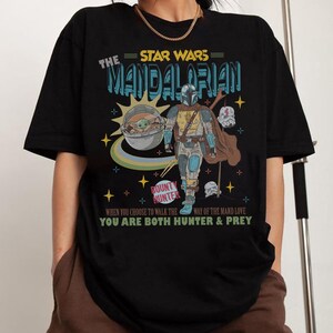 May include: Black t-shirt featuring a graphic of The Mandalorian from Star Wars. The design includes the text "The Mandalorian" and "Bounty Hunter", along with an image of the character and Grogu. The shirt also has the text "When you choose to walk the way of the Mand love, you are both hunter & prey."