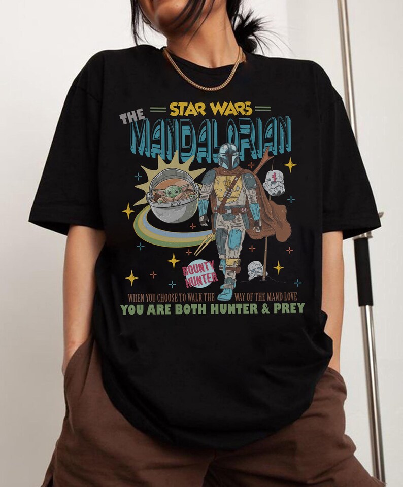 May include: Black t-shirt featuring a graphic of The Mandalorian from Star Wars. The design includes the text "The Mandalorian" and "Bounty Hunter", along with an image of the character and Grogu. The shirt also has the text "When you choose to walk the way of the Mand love, you are both hunter & prey."