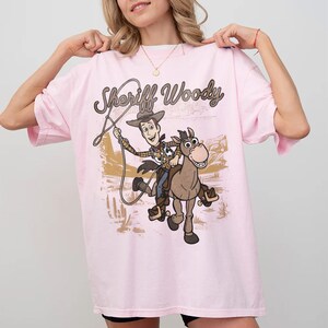 May include: A light pink t-shirt featuring a graphic print of Sheriff Woody riding Bullseye the horse. The design includes the text Sheriff Woody in a brown cursive font, with a desert landscape background featuring cacti and mountains.
