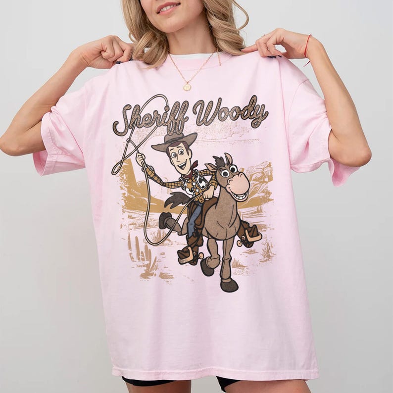 May include: A light pink t-shirt featuring a graphic print of Sheriff Woody riding Bullseye the horse. The design includes the text Sheriff Woody in a brown cursive font, with a desert landscape background featuring cacti and mountains.