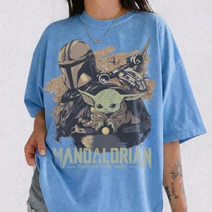 May include: Light blue t-shirt featuring a graphic of The Mandalorian, Grogu, and a spaceship. The design includes the text "MANDALORIAN - THIS IS THE WAY" in a distressed, vintage style. The shirt has short sleeves and a crew neck.
