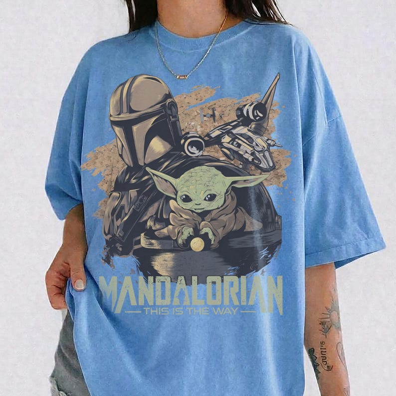 May include: Light blue t-shirt featuring a graphic of The Mandalorian, Grogu, and a spaceship. The design includes the text "MANDALORIAN - THIS IS THE WAY" in a distressed, vintage style. The shirt has short sleeves and a crew neck.