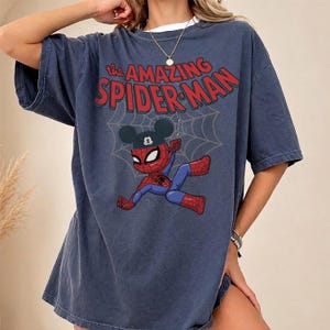 May include: A navy blue t-shirt featuring the text "The Amazing Spider-Man" in red. The graphic shows a cartoon Spider-Man wearing a Mickey Mouse hat, hanging from a web. The shirt has short sleeves and a crew neck.