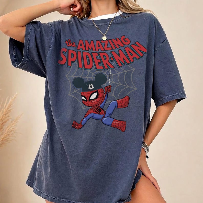 May include: A navy blue t-shirt featuring the text "The Amazing Spider-Man" in red. The graphic shows a cartoon Spider-Man wearing a Mickey Mouse hat, hanging from a web. The shirt has short sleeves and a crew neck.