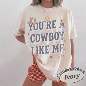 May include: An ivory short-sleeve t-shirt featuring the blue text "YOU'RE A COWBOY LIKE ME" printed on the front. The design includes illustrations of brown cowboy boots, a yellow ball, a sheriff star, blue clouds, red stars, and a rocket ship.