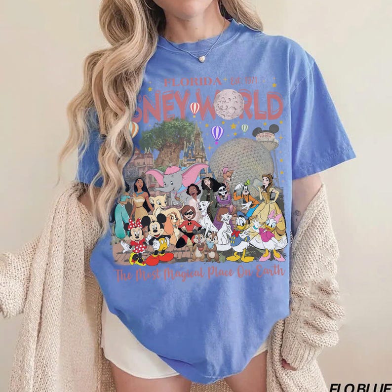 May include: A blue t-shirt featuring a colorful graphic of Disney World characters and landmarks. The text "Florida's Disney World" and "The Most Magical Place On Earth" are printed on the shirt. The shirt has short sleeves and a crew neck.