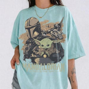 May include: A light blue t-shirt featuring a graphic of The Mandalorian, Grogu, and a spaceship. The design includes the text "MANDALORIAN" and "THIS IS THE WAY" in a light green color. The artwork is set against a sandy background.