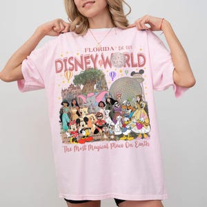 May include: Pink t-shirt with a Disney World graphic. The design includes the text "Florida Est. 1971" and "Disney World" in a retro font, along with illustrations of Disney characters and landmarks. The phrase "The Most Magical Place On Earth" is also included.