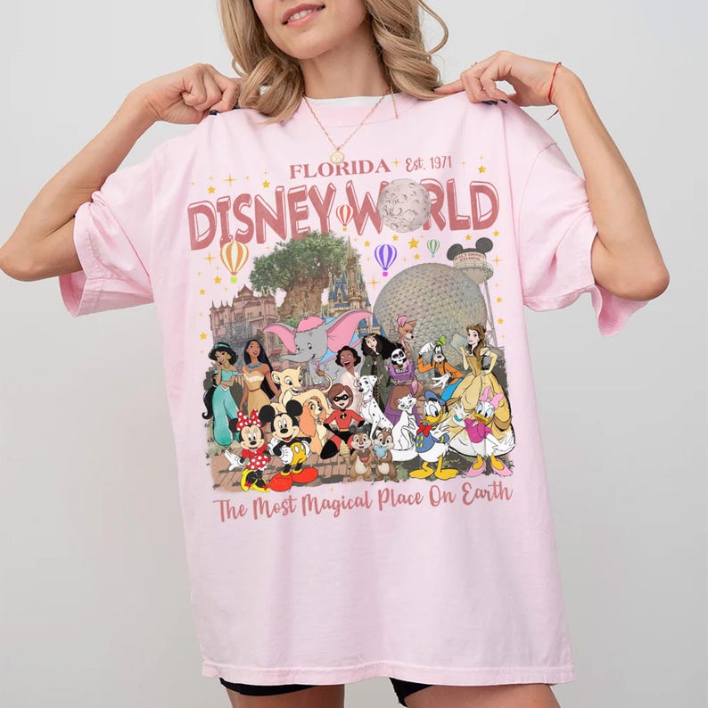 May include: Pink t-shirt with a Disney World graphic. The design includes the text "Florida Est. 1971" and "Disney World" in a retro font, along with illustrations of Disney characters and landmarks. The phrase "The Most Magical Place On Earth" is also included.