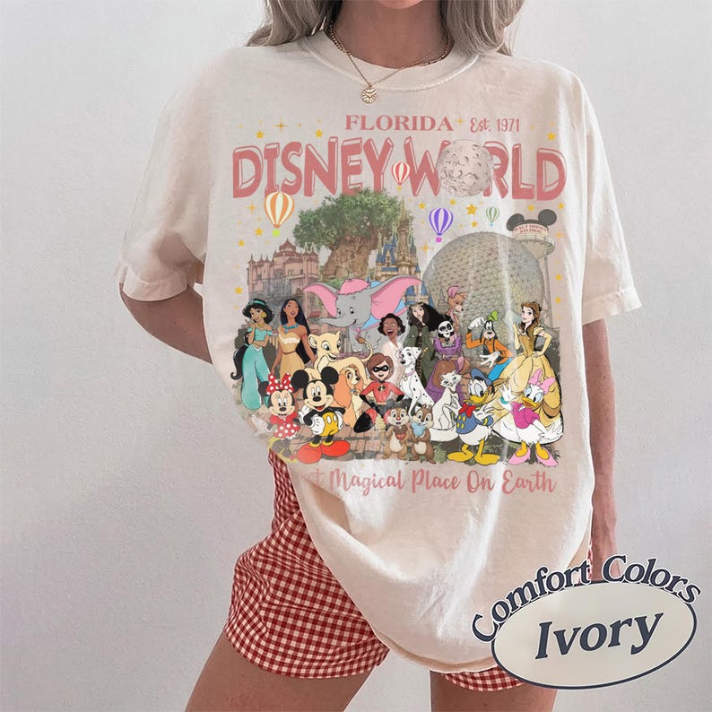May include: Ivory-colored t-shirt featuring a graphic of Disney World in Florida. The design includes the words "Disney World" and "Magical Place On Earth" with various Disney characters and park icons. The shirt is a Comfort Colors brand.