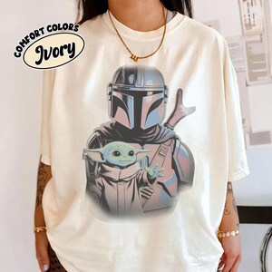 May include: An ivory t-shirt featuring a graphic print of a helmeted armored character standing behind a small green creature with large ears. The text COMFORT COLORS IVORY is printed in a black oval in the upper left corner of the shirt.