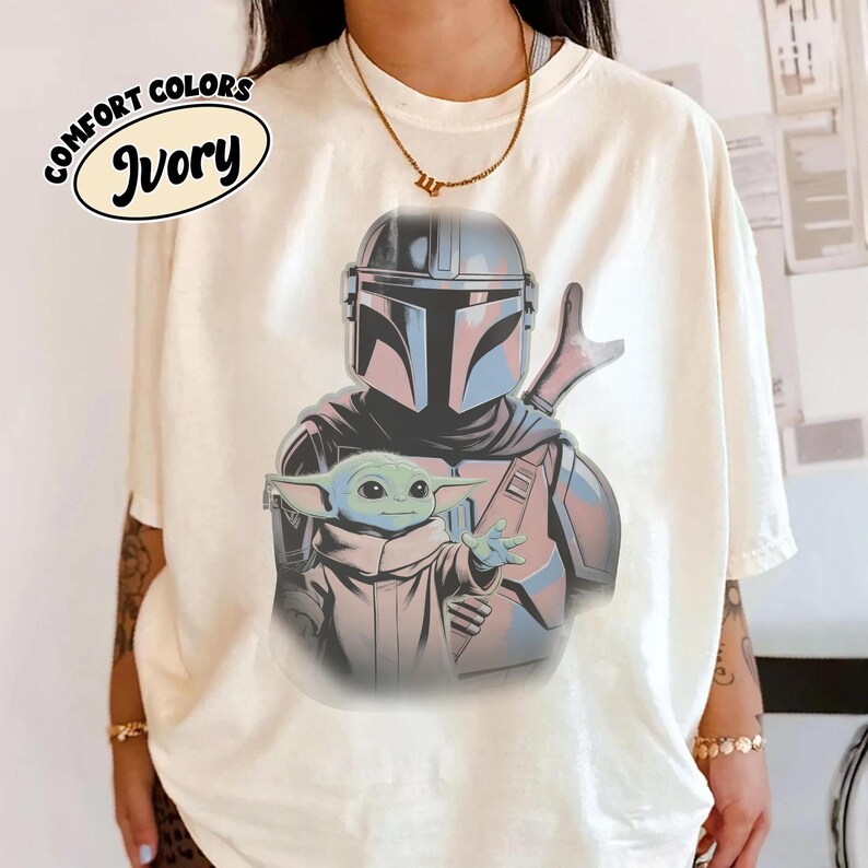 May include: An ivory t-shirt featuring a graphic print of a helmeted armored character standing behind a small green creature with large ears. The text COMFORT COLORS IVORY is printed in a black oval in the upper left corner of the shirt.