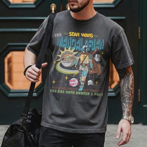May include: Dark gray t-shirt featuring a graphic of The Mandalorian, Grogu, and other characters from the Star Wars series. The design includes the text "The Mandalorian" and "Bounty Hunter".
