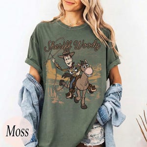 May include: A moss green crew neck t-shirt featuring a graphic print of Sheriff Woody riding Bullseye. The text Sheriff Woody is printed above the characters in a brown script font. The design includes a lasso and a desert landscape background.