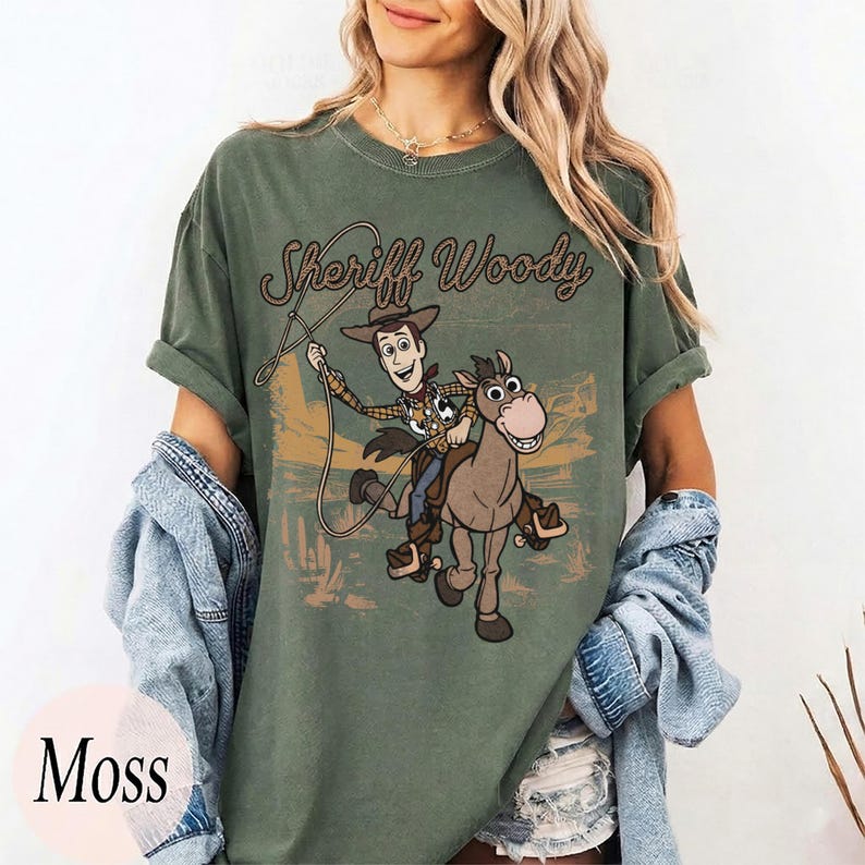 May include: A moss green crew neck t-shirt featuring a graphic print of Sheriff Woody riding Bullseye. The text Sheriff Woody is printed above the characters in a brown script font. The design includes a lasso and a desert landscape background.