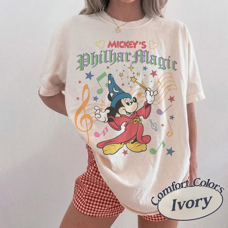May include: Ivory-colored t-shirt featuring Mickey Mouse as a sorcerer with the text "Mickey's PhilharMagic." The design includes musical notes, stars, and a wand. The shirt is labeled "Comfort Colors Ivory."