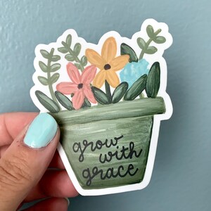 May include: A green flower pot with a white outline, filled with flowers in shades of pink, yellow, and blue. The pot has the text "grow with grace" written on it.