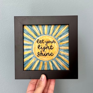 May include: Square framed artwork featuring a yellow sunburst design with the words "let your light shine" in black script. The sun is centered on a blue and yellow background, and the frame is black.