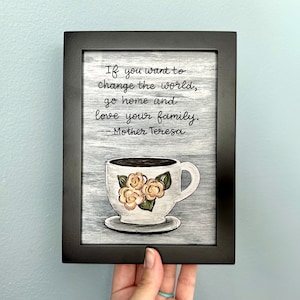 May include: A framed black and white sign with a quote by Mother Teresa: "If you want to change the world, go home and love your family." The sign features a painted image of a white coffee cup with yellow flowers.
