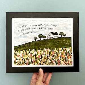 May include: A framed print with a white house on a green hill with a field of flowers in the foreground. The text reads "I still remember the days I prayed for the things I have now."