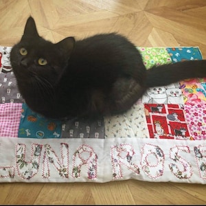 May include: A black cat with yellow eyes is lying on a patchwork quilt with the name "Luna Rose" embroidered on it.