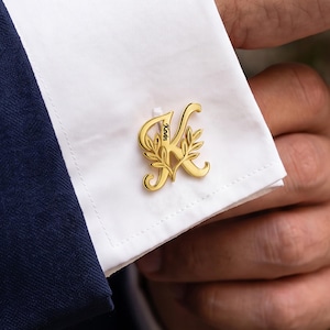 Op de afbeelding: Een goudkleurige manchetknop met een gestileerde letter K met een geïntegreerd bladmotief en de naam Kevin verticaal gegraveerd langs de centrale lijn van de letter, bevestigd aan een wit overhemdmanchet met een donkerblauwe mouw.