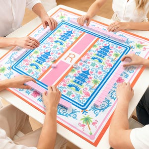 May include: A square Mahjong game mat with a pink and blue floral design, featuring pagodas, flamingos, and palm trees. The mat is surrounded by pink tiles, with players' hands visible. The central design includes the letter "R".