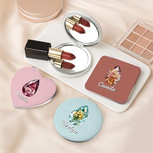 Könnte beinhalten: Kompakte Kosmetikspiegel in Rosa, Blau und Braun mit floralen Designs und Namen. Ein herzförmiger, ein runder und ein quadratischer Spiegel sind abgebildet. Ein Lippenstift und eine Lidschattenpalette sind ebenfalls zu sehen.