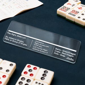 May include: A clear acrylic game guide with white text, including game rules and instructions. The guide is surrounded by ivory-colored game tiles with red and black markings, and a sheet of paper with additional game instructions.